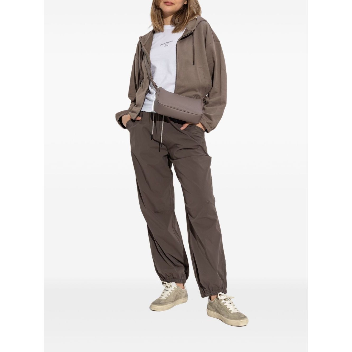 Woman in casual gray hoodie and pants outfit, wearing white sneakers, standing against plain background.