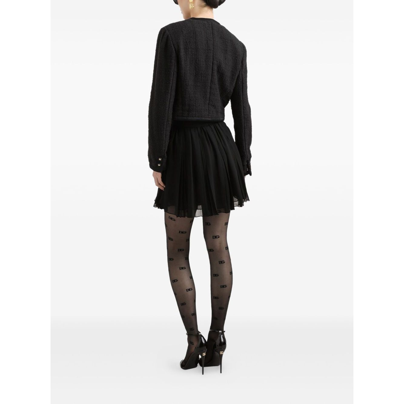 Back view of model wearing Dolce & Gabbana Rachel Tweed Jacket, black crew neck, buttoned cuffs, with a pleated skirt and patterned tights.